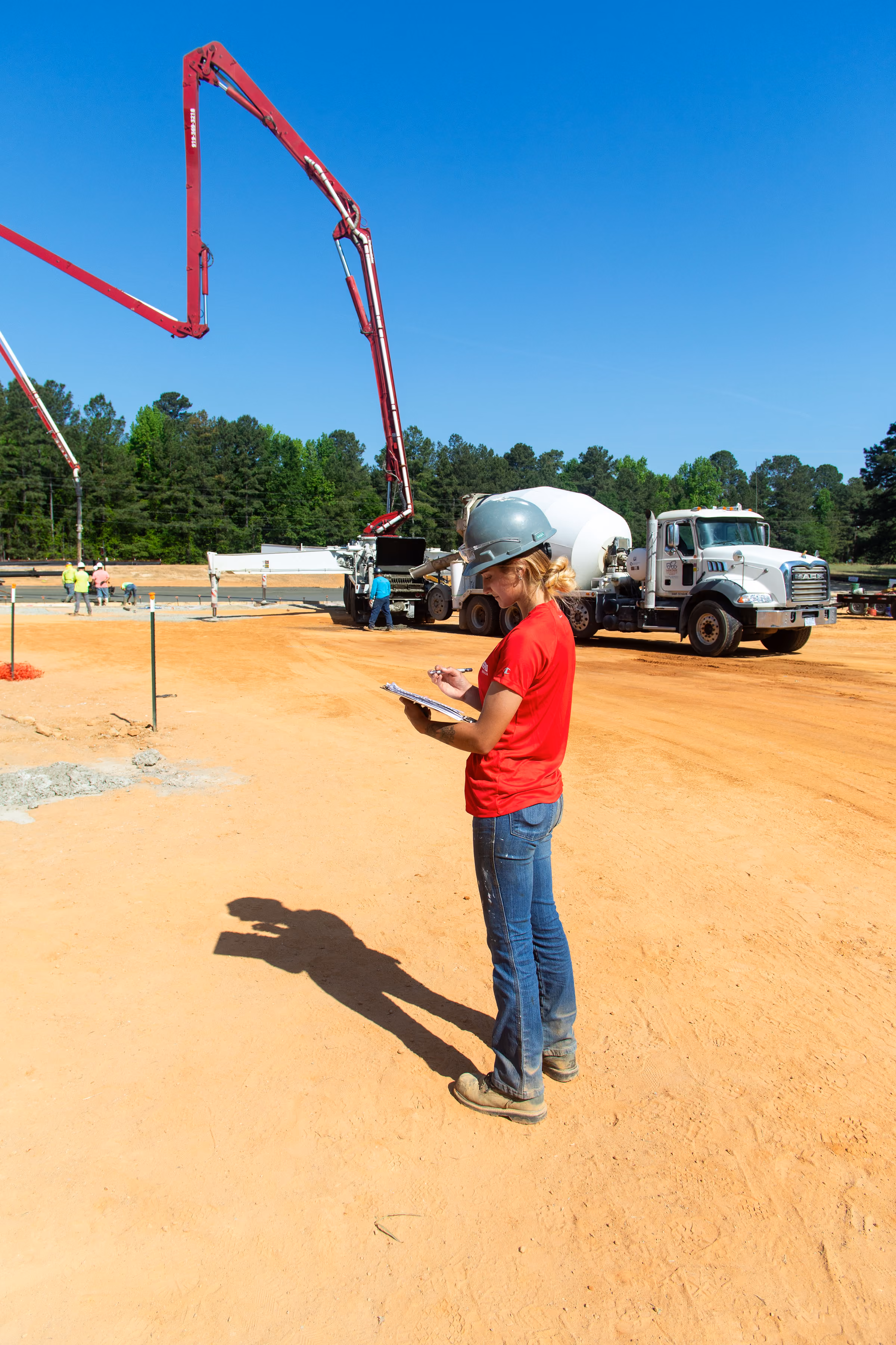 Construction worker in red shirt and helmet writes on clipboard at dirt site with cement truck and boom pump.