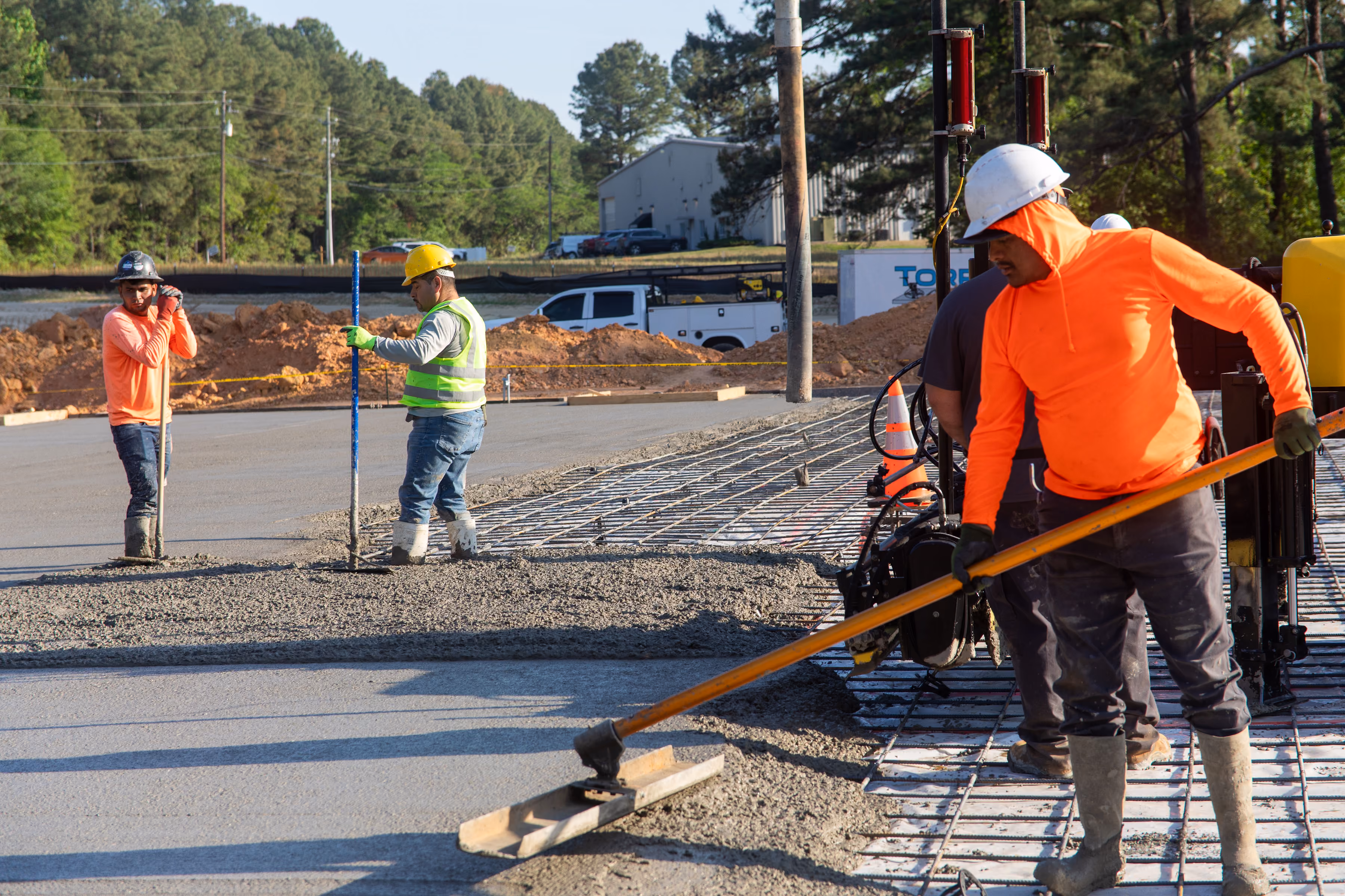 Construction workers smoothing wet concrete on a road with tools and safety gear under daylight.