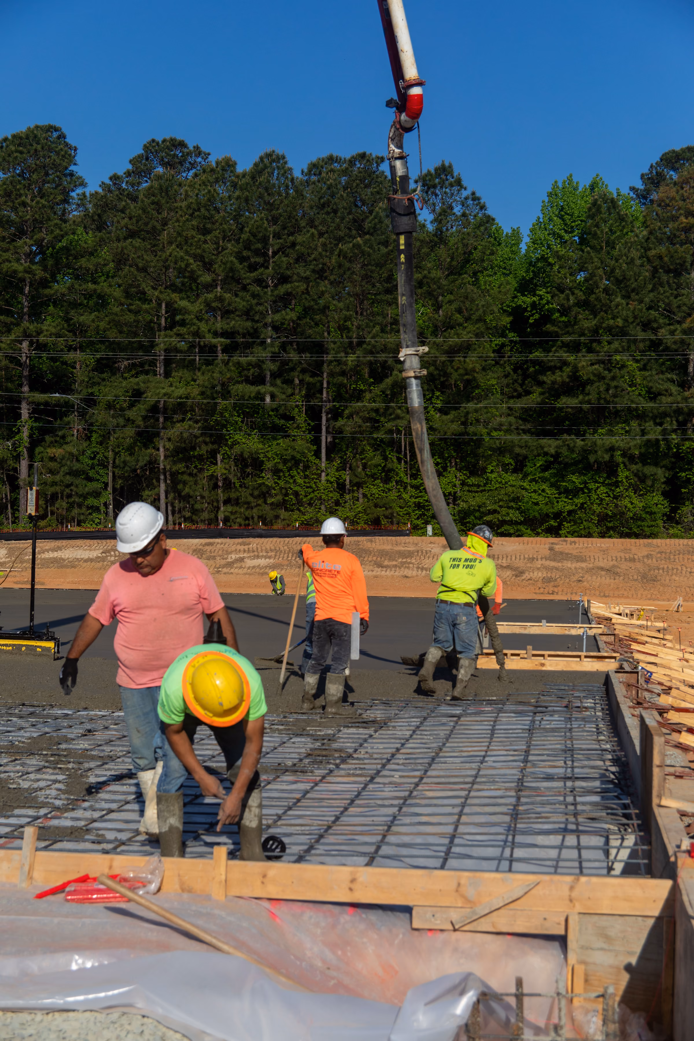 Construction workers pouring and leveling concrete on a large outdoor site with trees in the background.