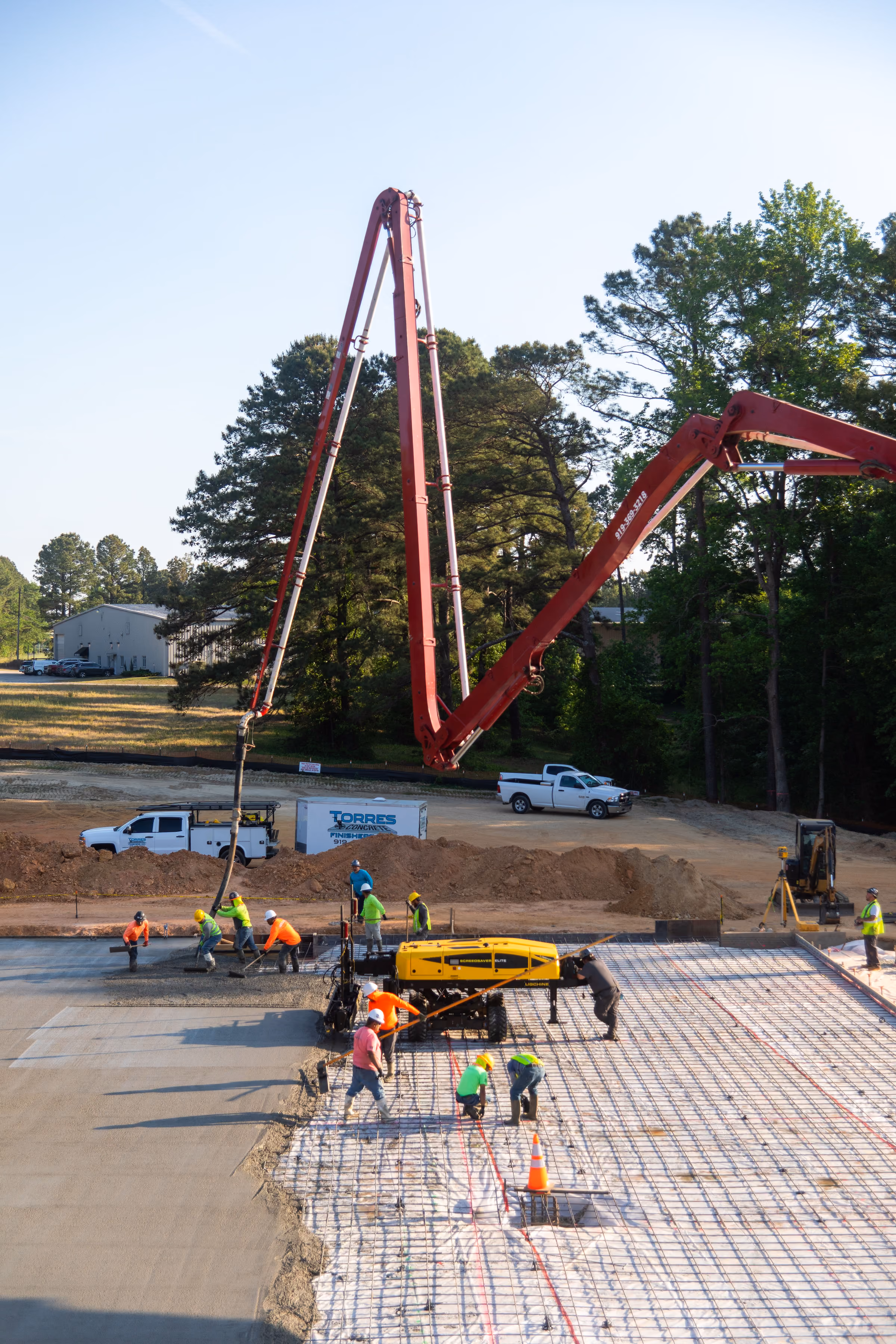 Construction workers pouring concrete with machinery on a large outdoor site with trees in the background.