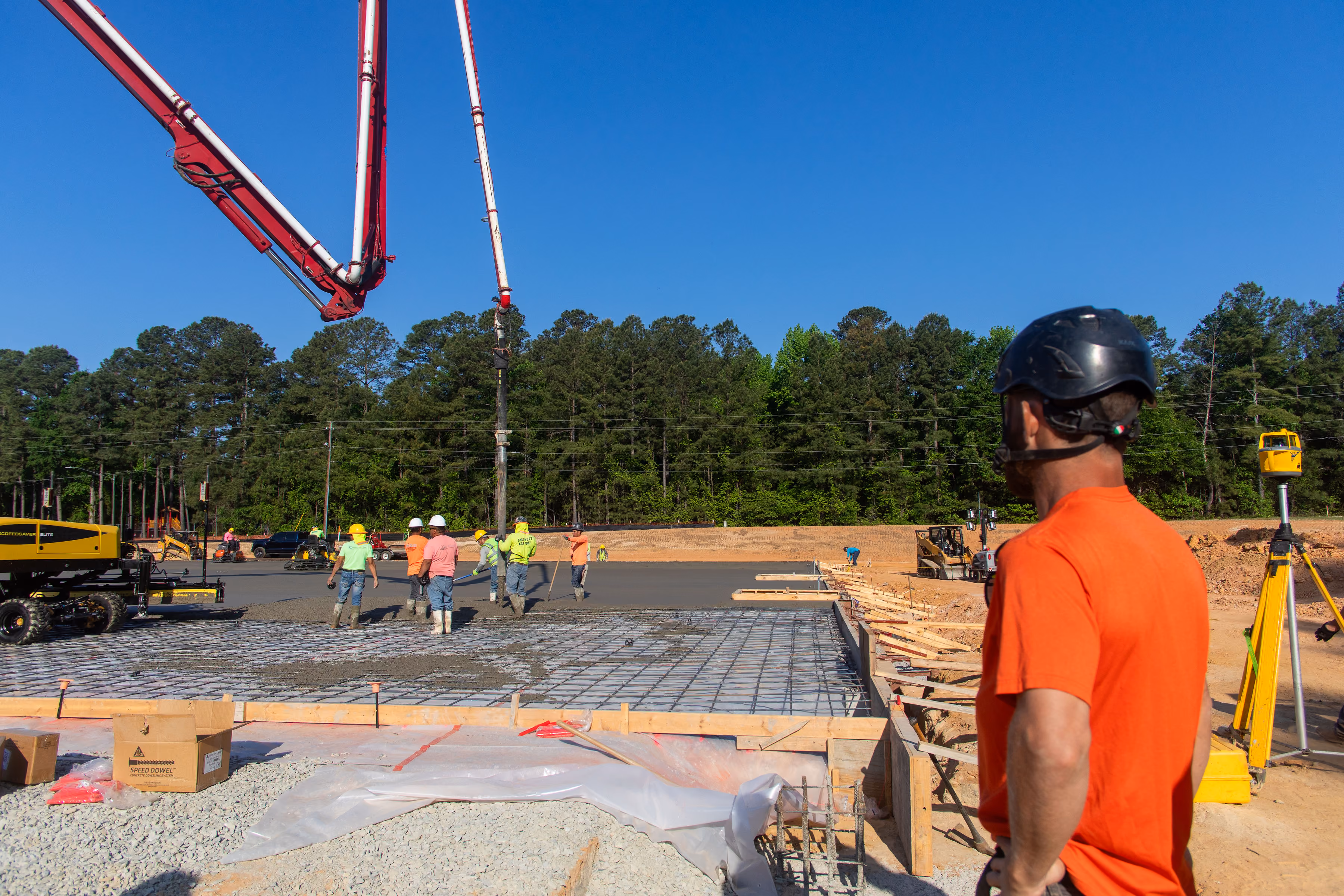 Workers pour concrete on foundation at sunny construction site with trees in background.