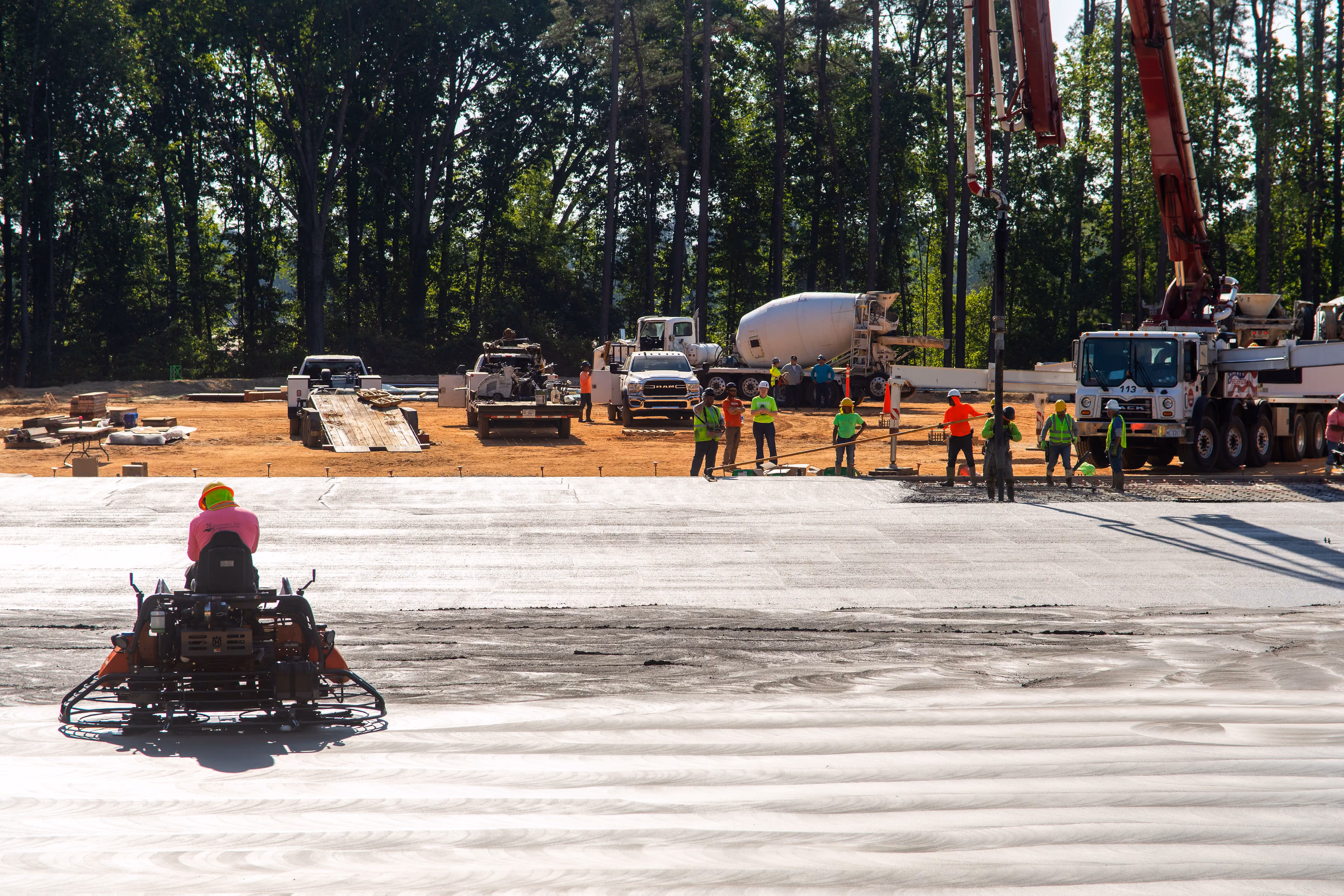 Workers laying and smoothing concrete on a large construction site with trucks and trees in background.