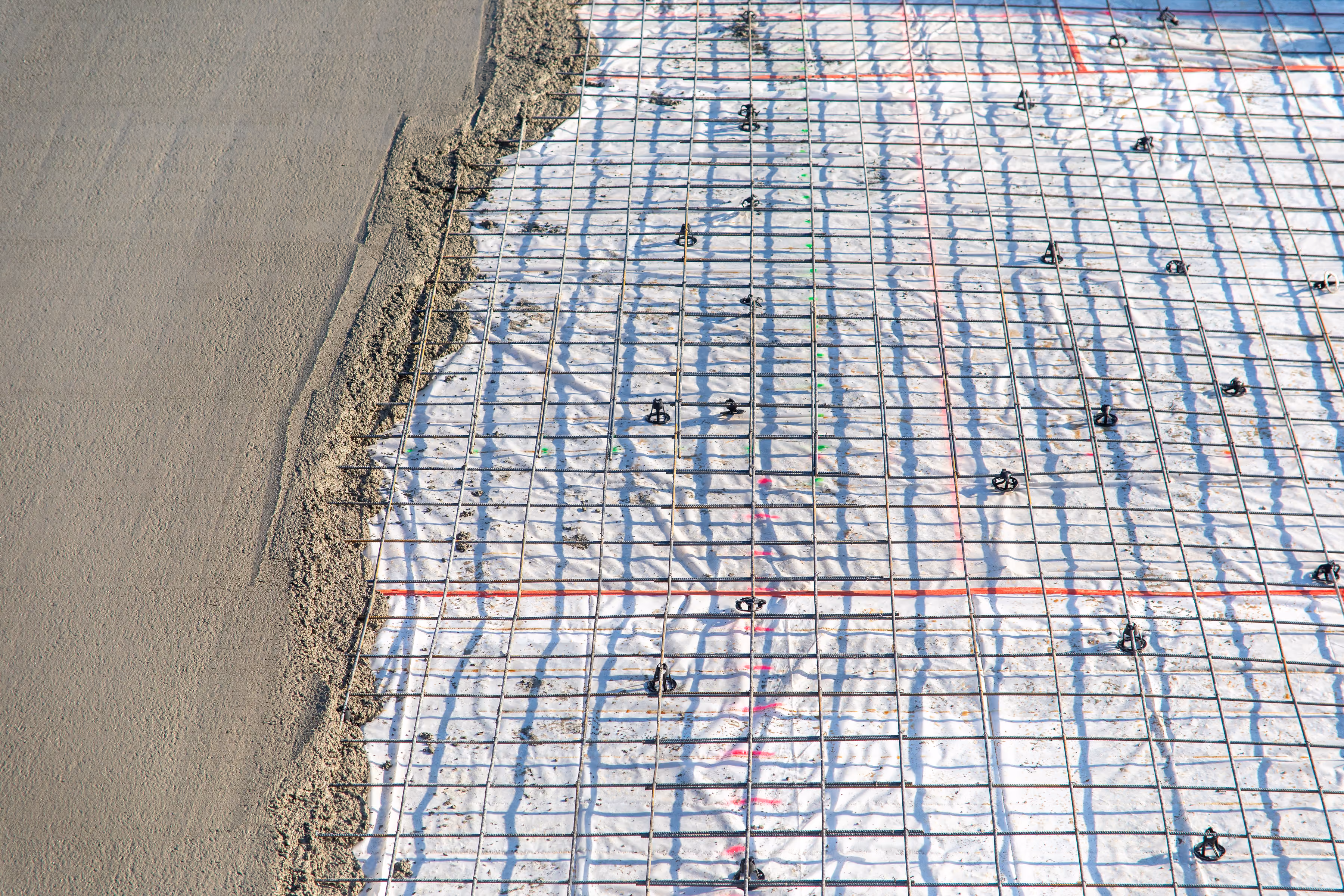 Construction site with wet concrete poured beside a grid of steel rebar on white plastic sheeting.