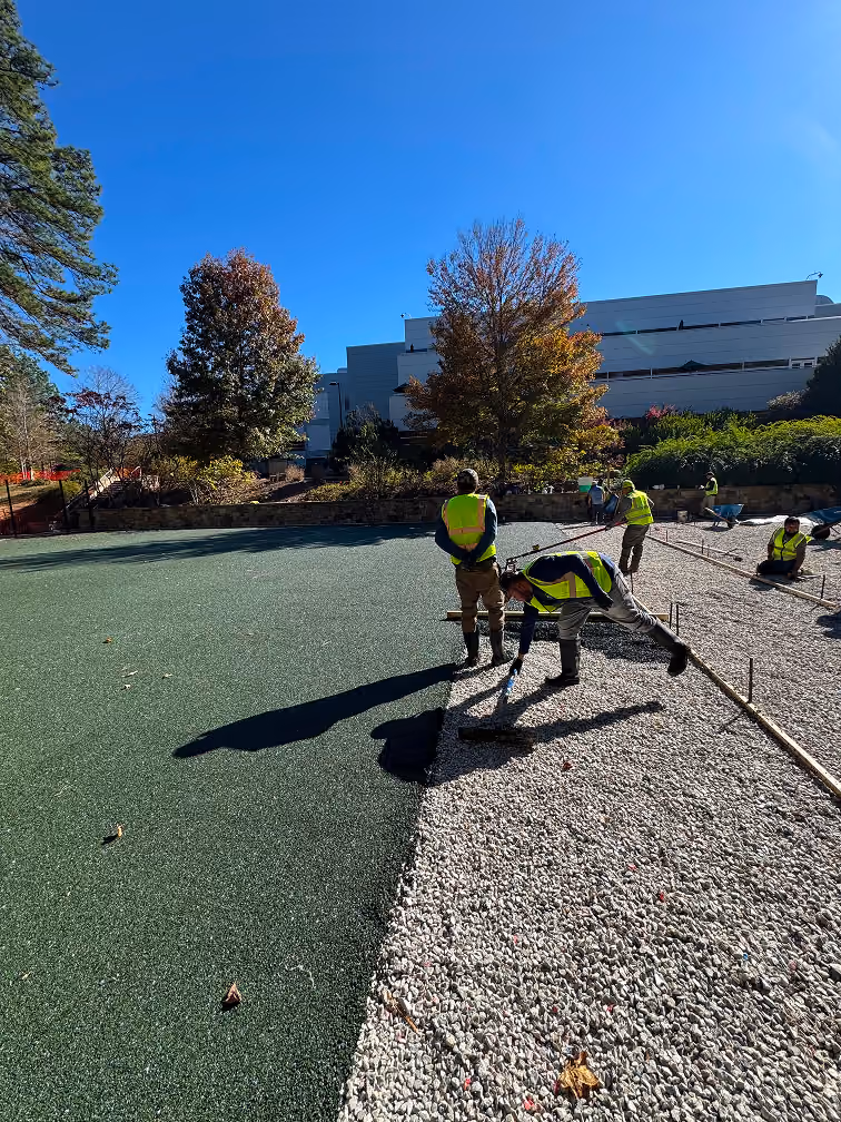 Workers in safety vests spreading green material on gravel surface outdoors on a sunny day.