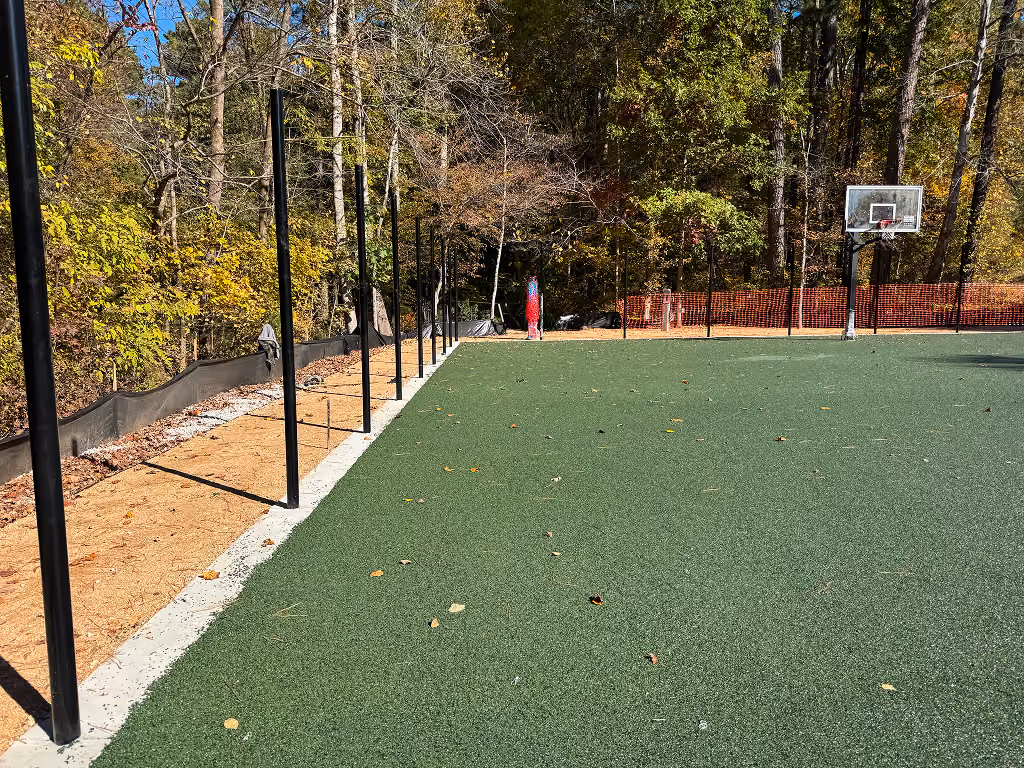 Outdoor basketball court with green turf, black poles along edge, and trees in background.