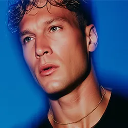 Close-up portrait of a young man with green eyes, curly dark hair, wearing a gold chain necklace and a black shirt against a blue background.