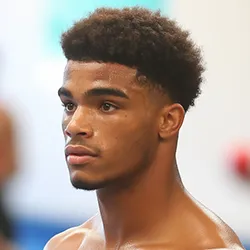 Close-up of a young man with short curly hair and a focused expression, looking slightly to the left against a blurred background.