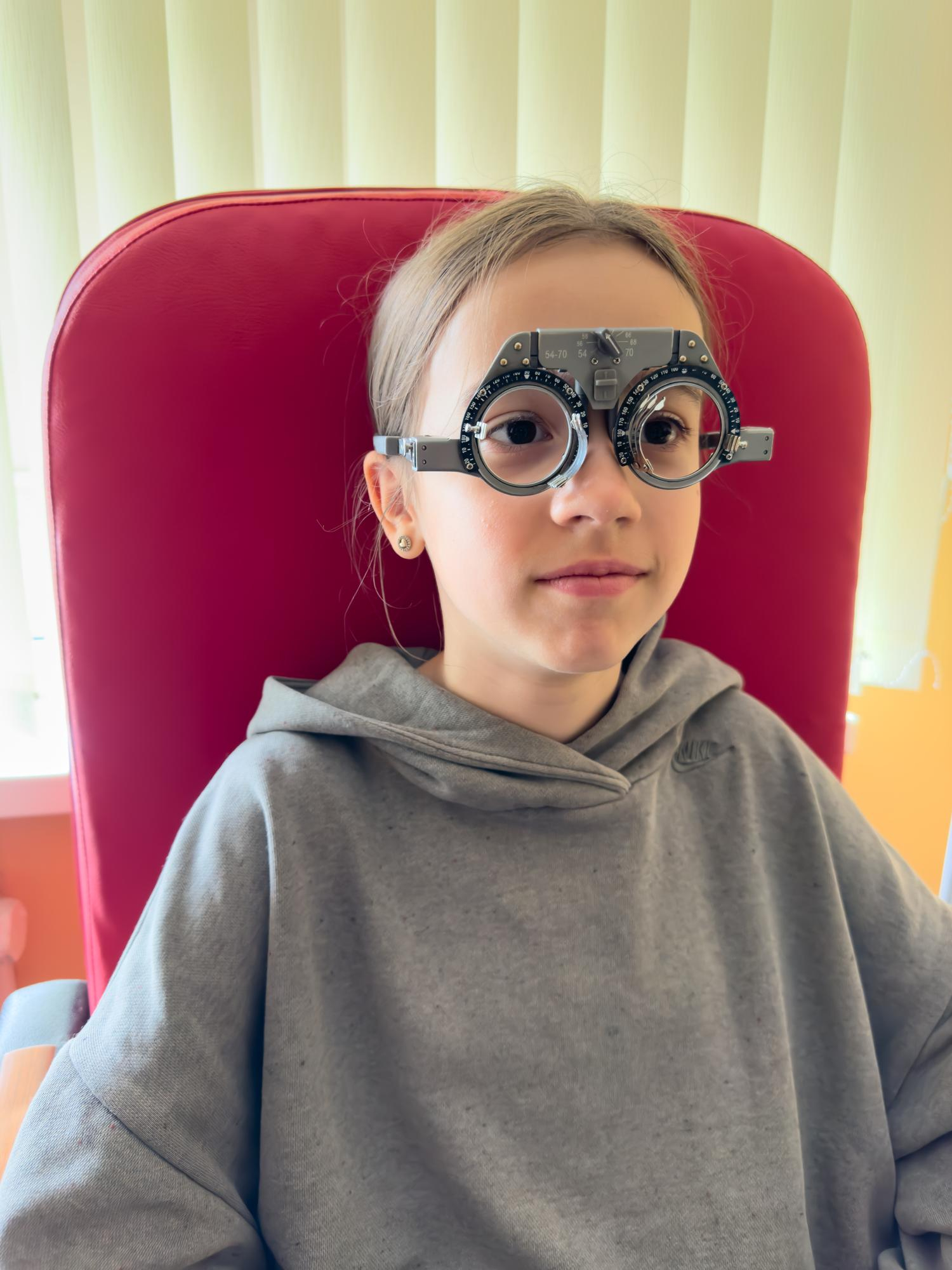 A young girl sitting in a red chair wearing a gray Nike hoodie and a black and silver trial frame used for eye exams.