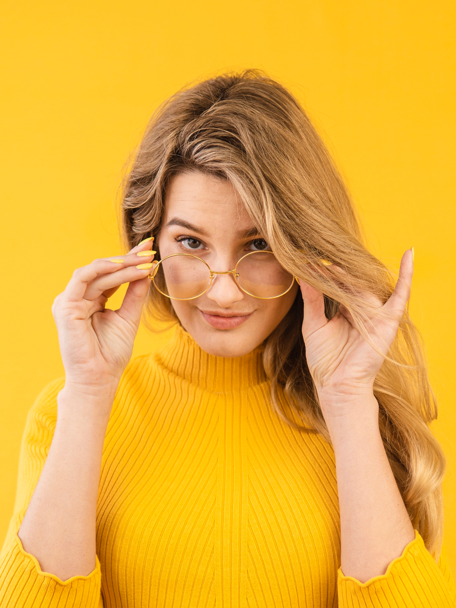 Young woman with long blonde hair wearing a yellow ribbed turtleneck sweater and gold-rimmed glasses, holding the glasses slightly away from her face while looking directly at the camera, against a solid yellow background.