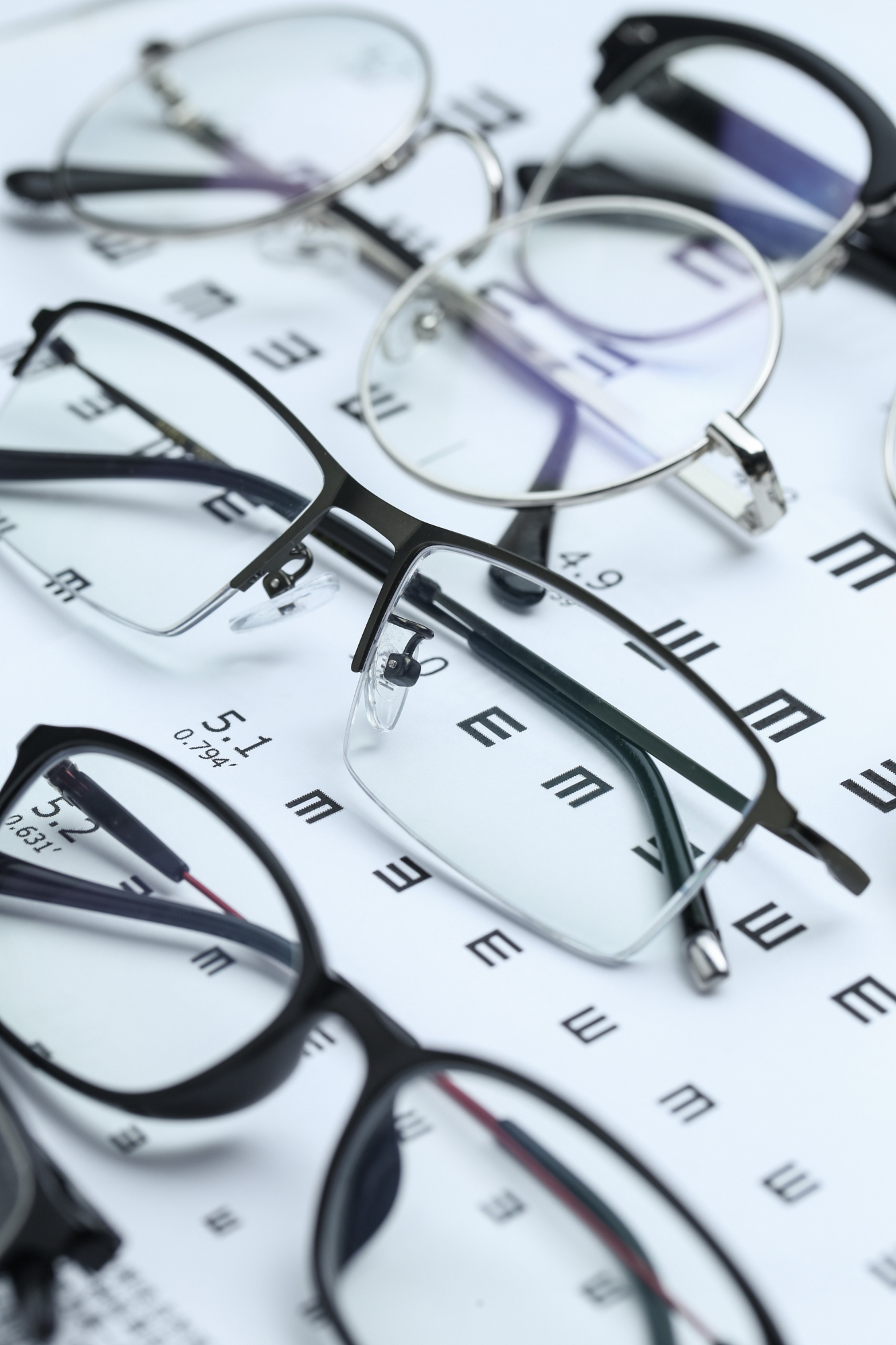 Close-up view of multiple pairs of eyeglasses with different frames resting on an eye chart with letters and numbers for vision testing.