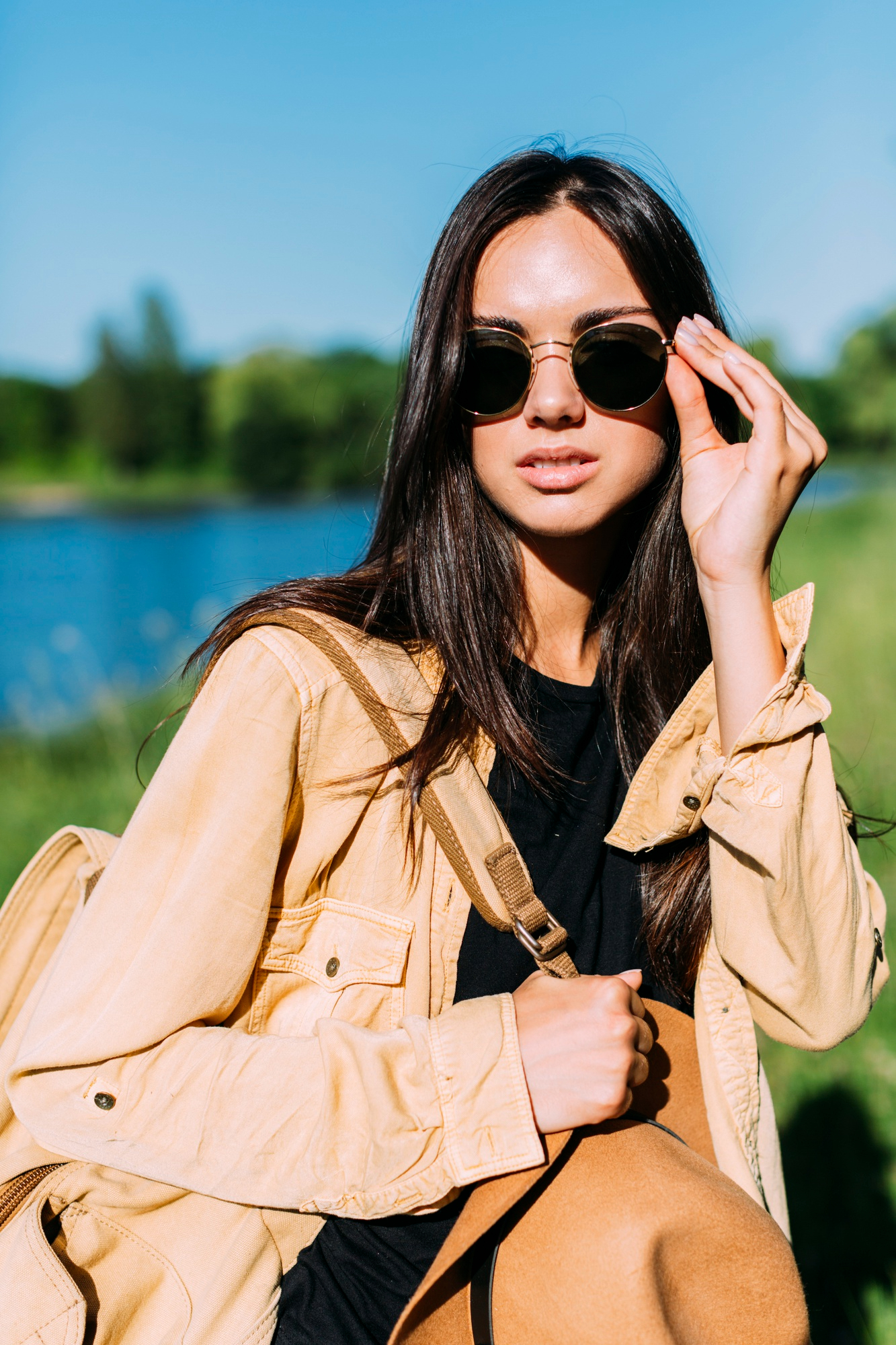 Young woman with long dark hair wearing round sunglasses, a beige jacket, and a black shirt, holding a brown hat and a backpack strap, standing outdoors near a body of water with green foliage in the background.