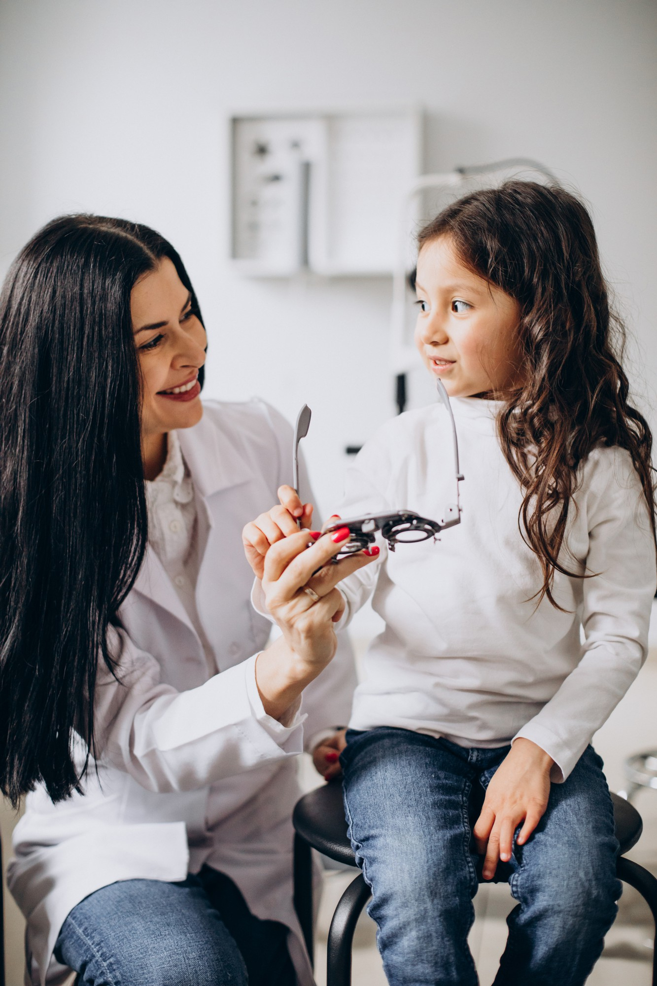 Female optometrist smiling and holding a trial frame while interacting with a young girl sitting on a stool in an eye exam room.