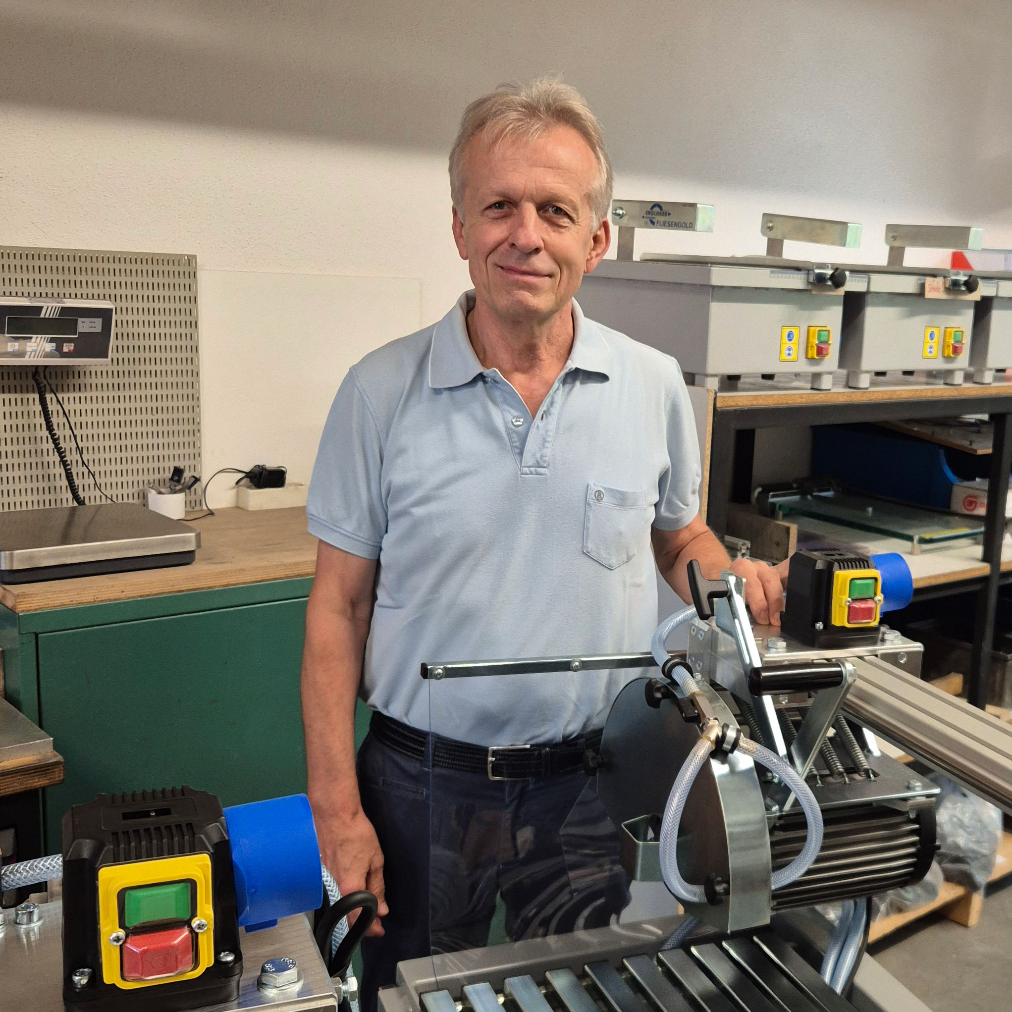 Älterer Mann in hellblauem Poloshirt steht in Werkstatt neben Metallmaschine mit Steuerknöpfen und Schläuchen.
