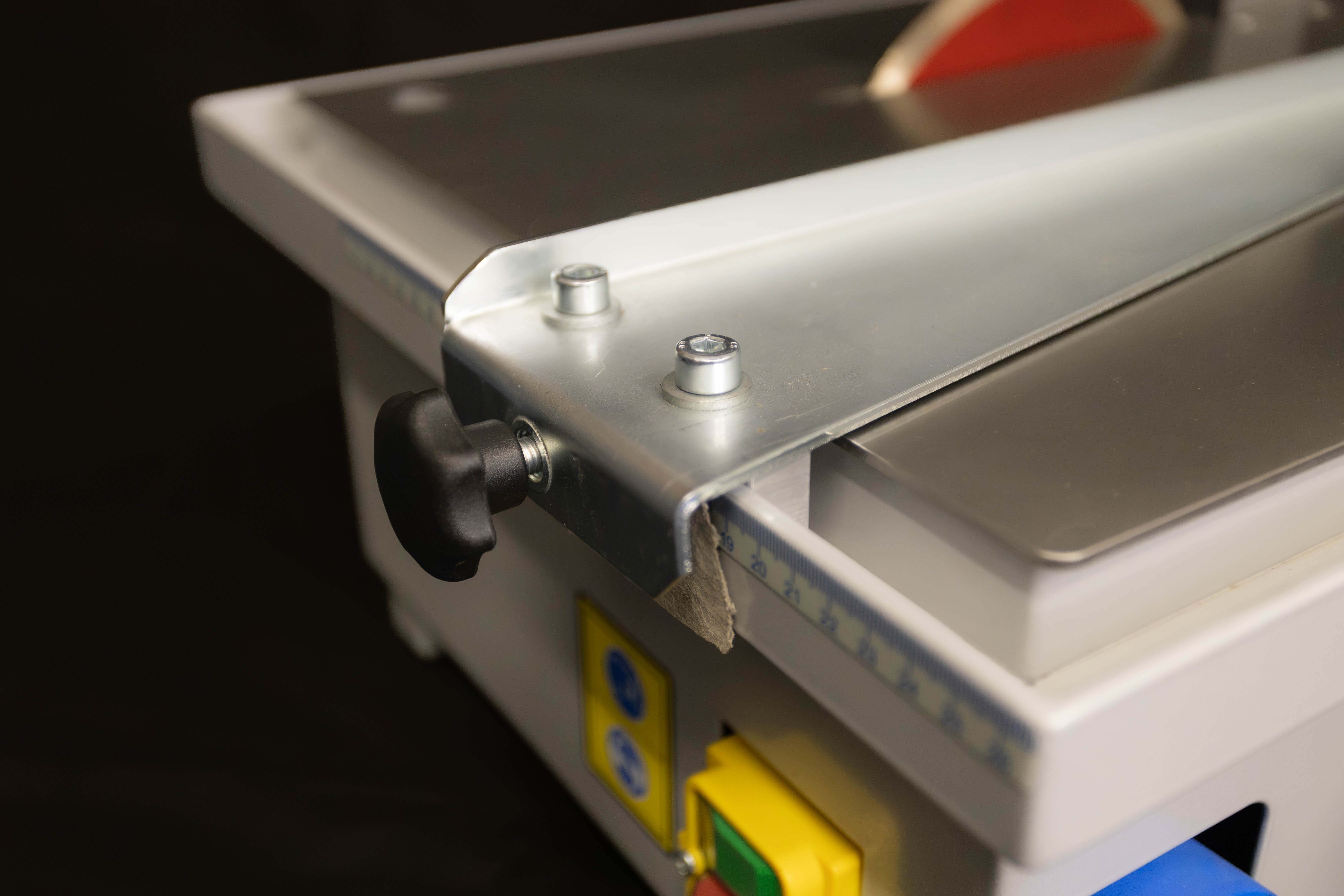 Nahaufnahme eines Metallhebers an einer elektrischen Tischsäge mit Messskala, schwarzem Drehgriff und gelbem Ein-/Ausschalter.