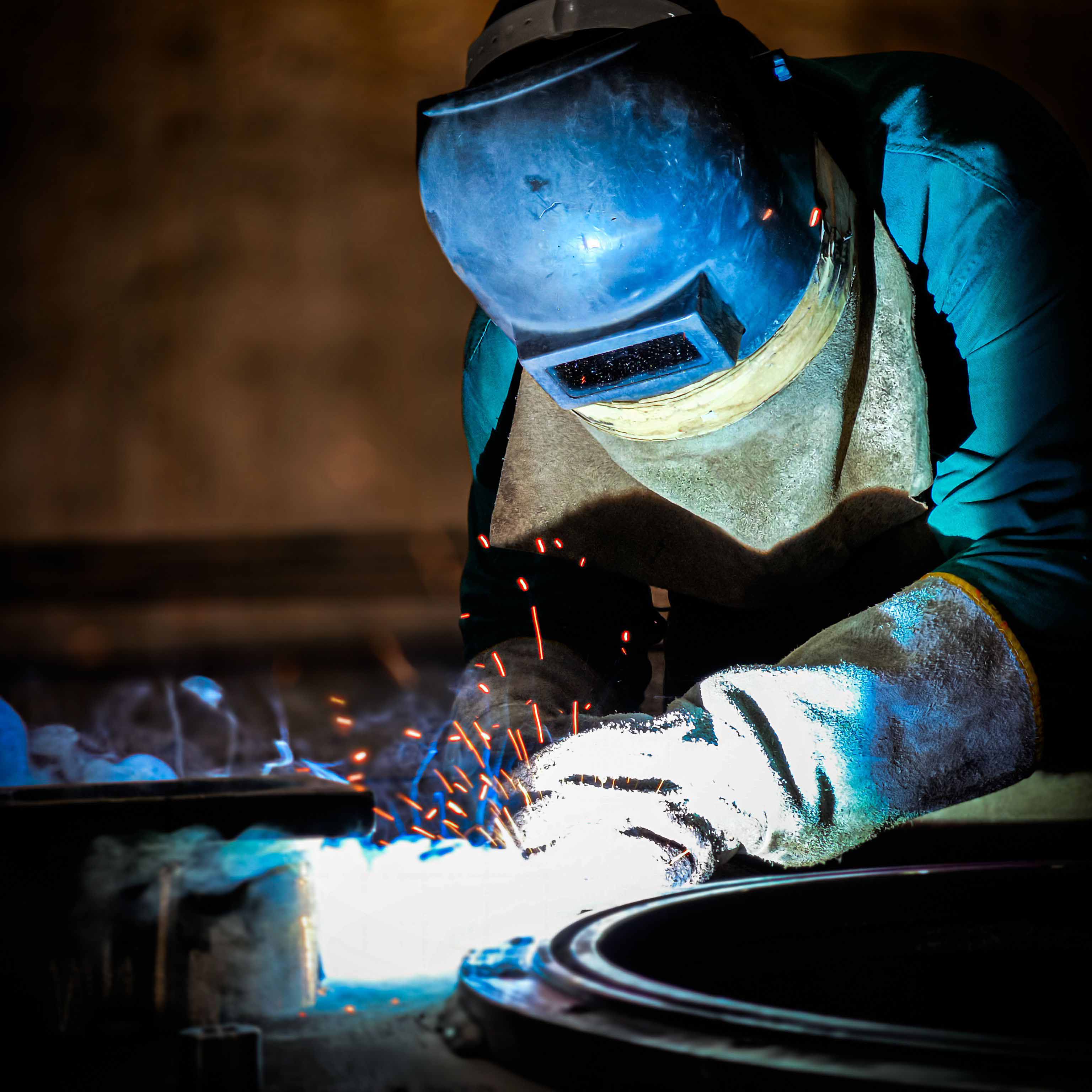 Schweißer mit Schutzhelm und Handschuhen arbeitet mit Funkenflug und blauem Licht beim Schweißen eines Metallteils.