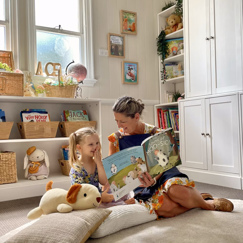 An educator reading a picture book with a young girl beside the cottage bookshelves.