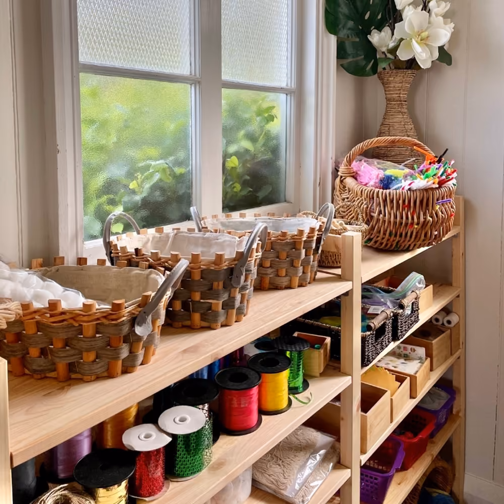 Open shelves of art materials in timber trays and woven baskets in the craft area.