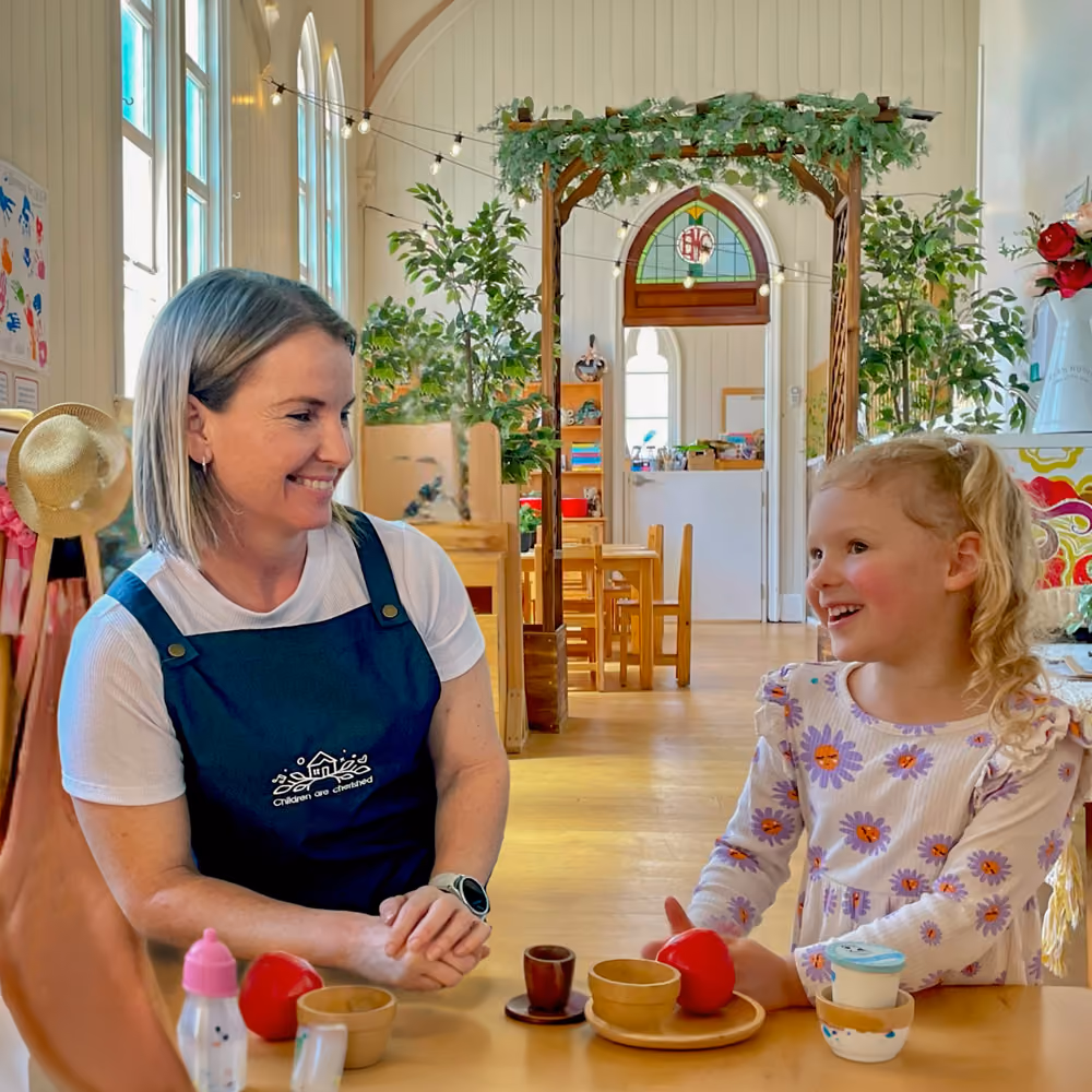 A Kindy Cottage Sandgate educator and a young girl smiling together in the playroom.