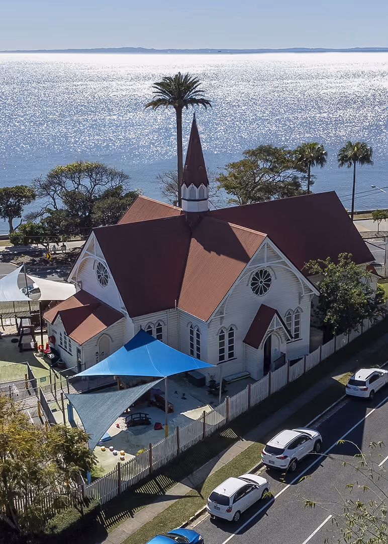 The red-roofed heritage building at Kindy Cottage Sandgate seen from above, surrounded by trees.