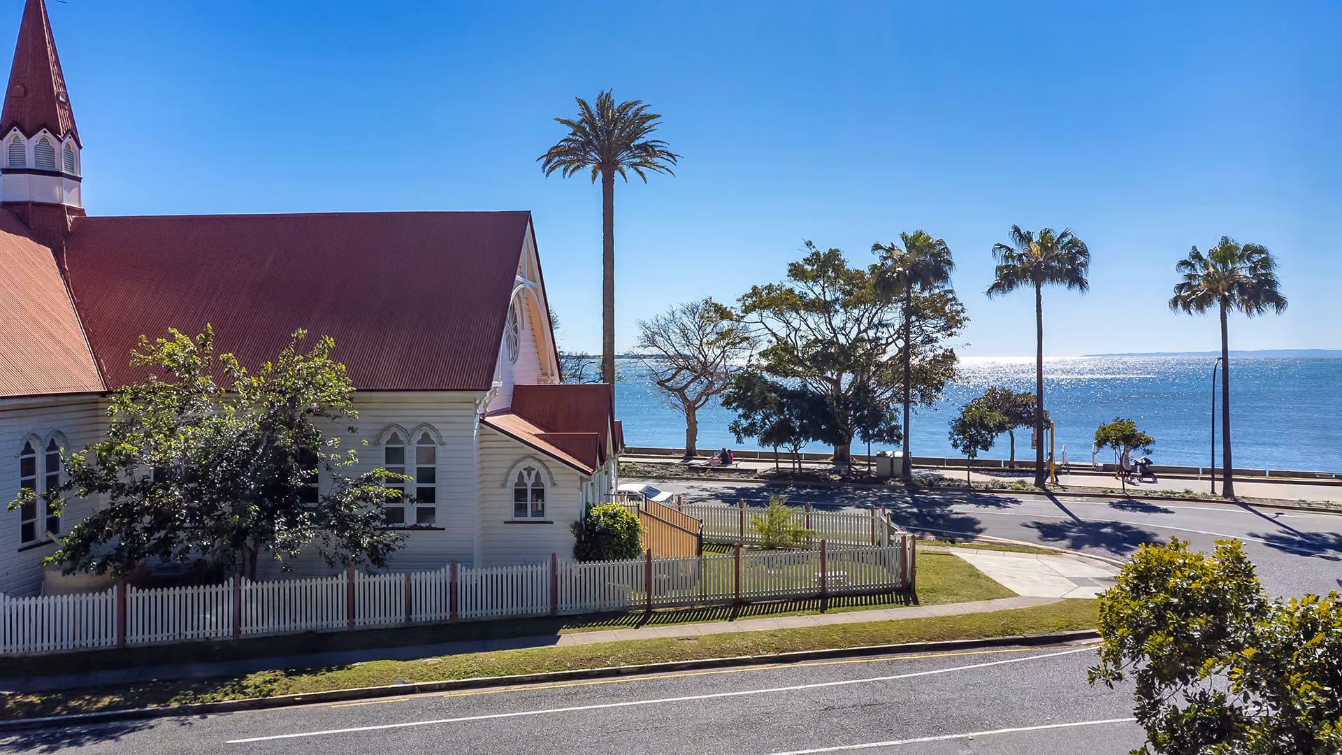 The heritage Kindy Cottage Sandgate building with its red roof, framed by palms near the bay.