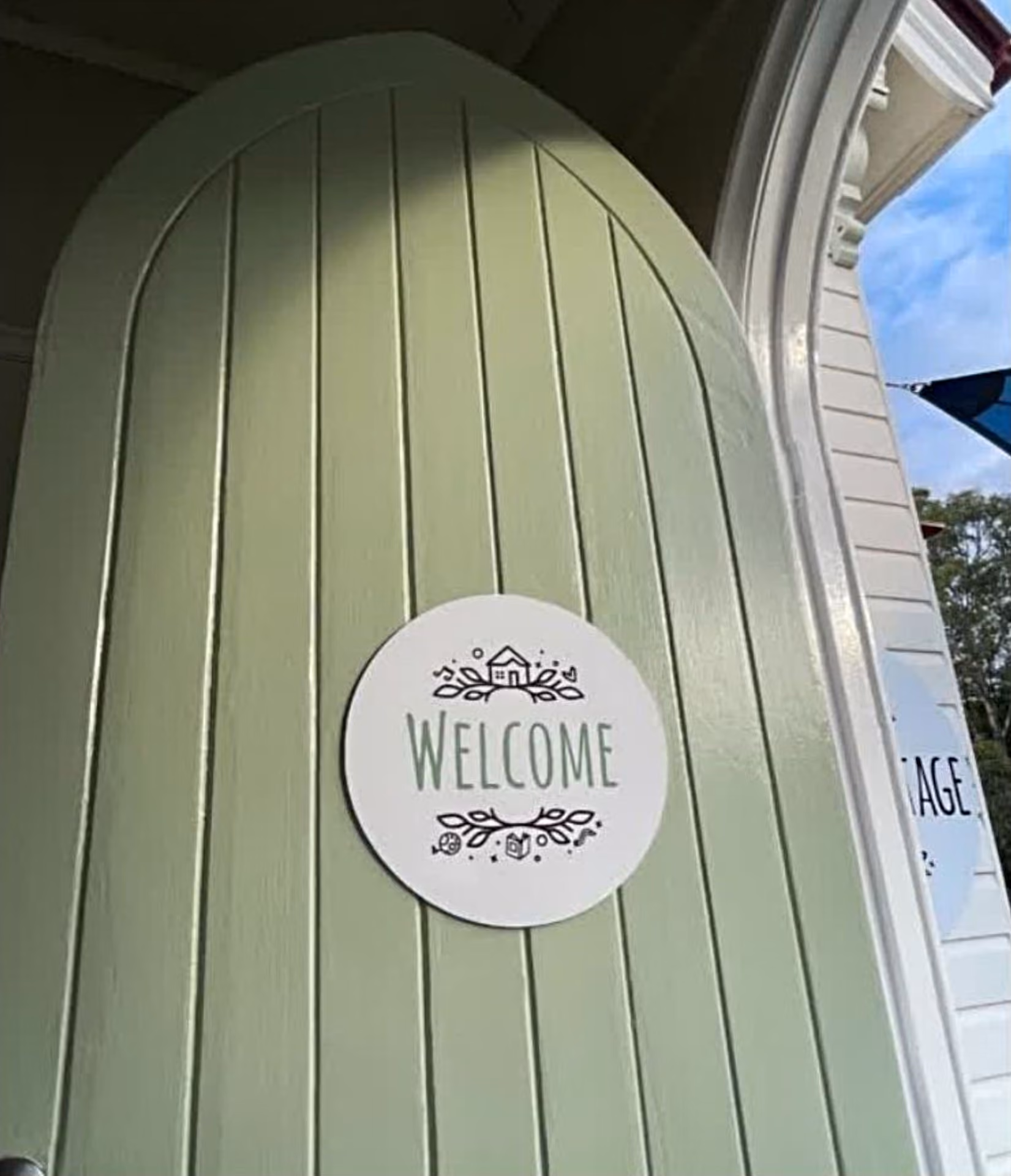 A round timber Welcome sign on the cottage's sage green weatherboard wall.