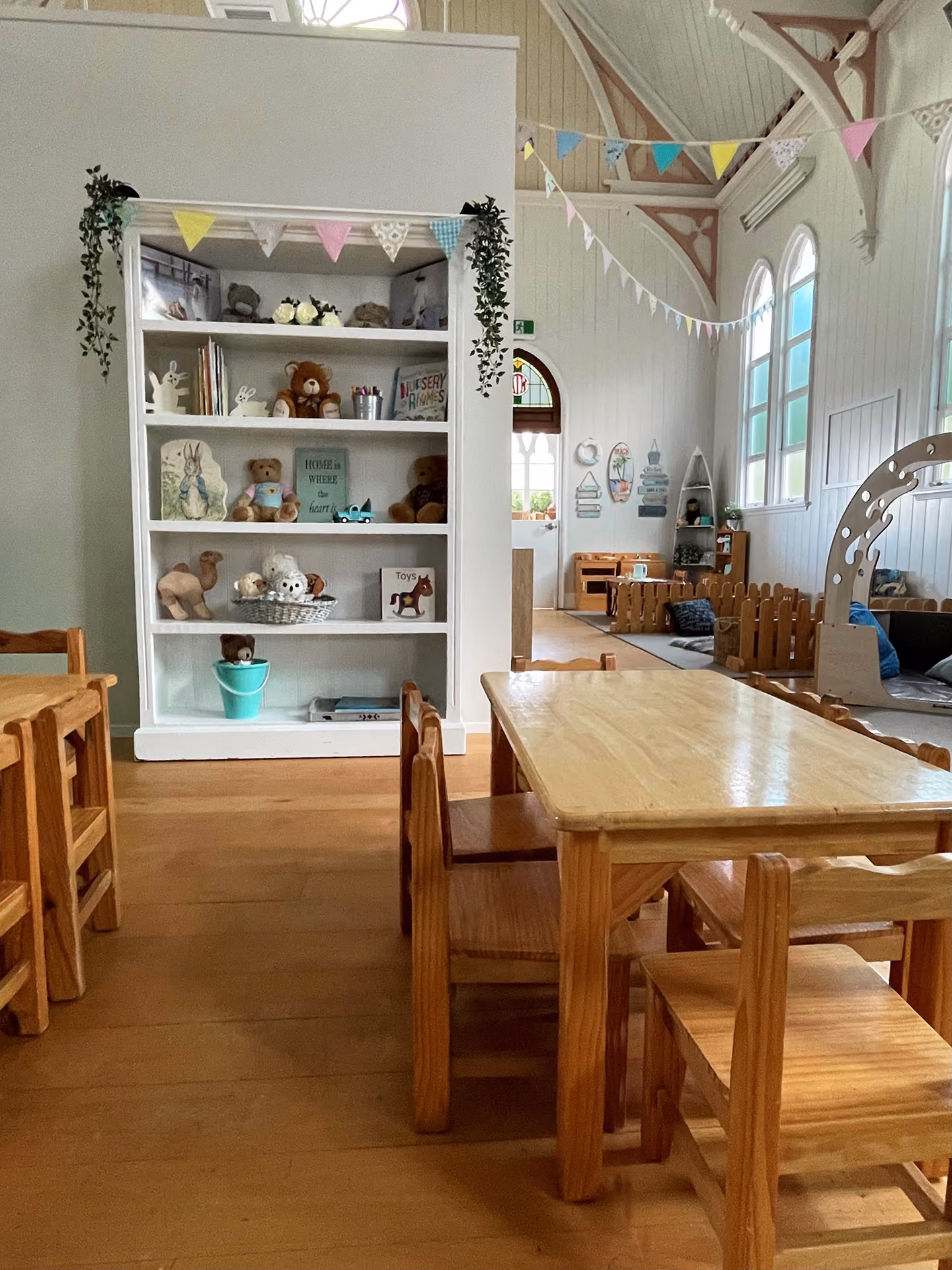 Children's playroom with wooden tables, chairs, toys on white shelves, and pastel bunting decorations.