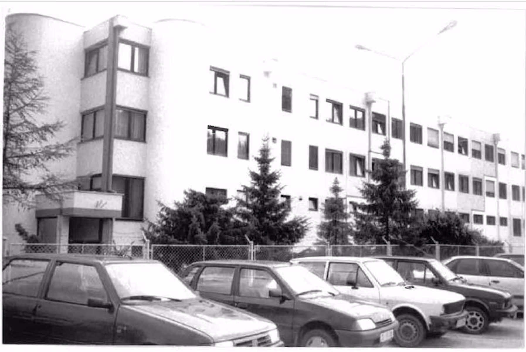 Black-and-white photograph of the three-storey toolshop building with a rounded stairwell tower, conifers and cars parked in front.