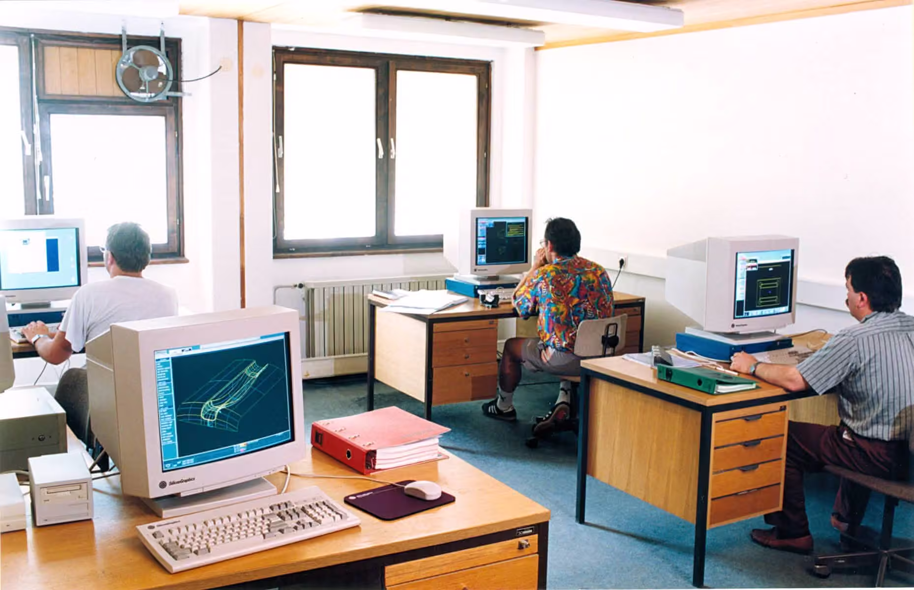 Three engineers at CRT computer workstations in the design office; the nearest screen shows a green CAD wireframe of a formed panel.