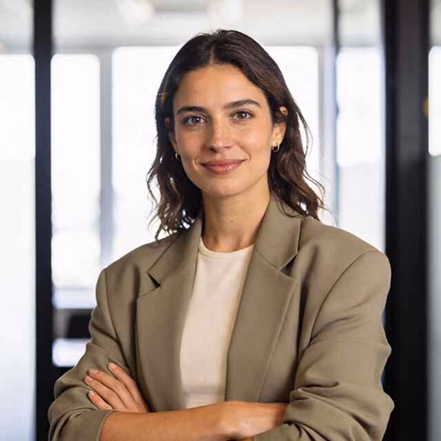 Confident professional woman with medium-length dark wavy hair, wearing a beige blazer and light-colored top, standing with arms crossed in a modern office environment.