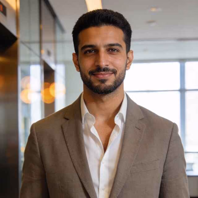 Confident man with dark hair and beard wearing a light brown suit jacket and white shirt, standing indoors with blurred window and lights in the background.