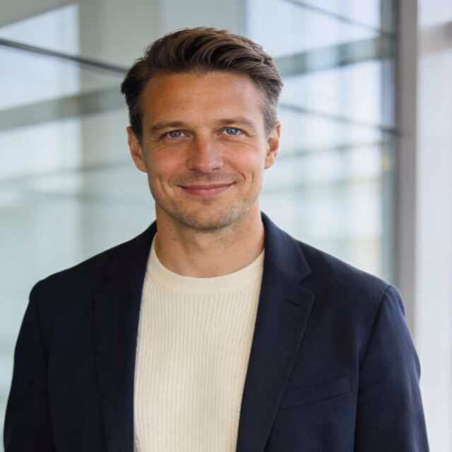 Smiling man with short brown hair and blue eyes wearing a black blazer over a cream sweater, standing in front of a glass window in an office setting.