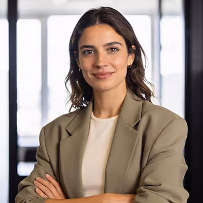Confident woman with dark wavy hair wearing a beige blazer and light-colored top, standing with arms crossed in a bright office space.