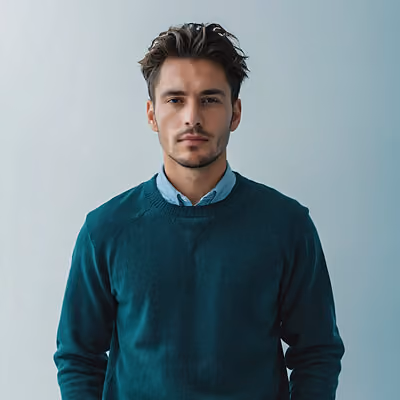 Portrait of a young man with short dark hair and light facial stubble, wearing a dark green sweater over a light blue collared shirt, standing against a plain light gray background.