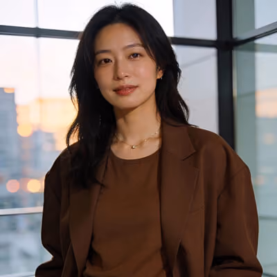 Young woman with long black hair wearing a brown blazer and matching top, standing indoors with large windows and city lights blurred in the background.