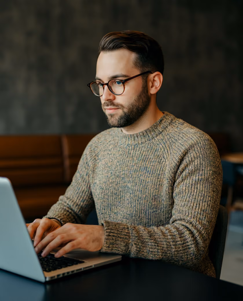 Young man with glasses and a beard wearing a knit sweater, typing on a laptop in a dimly lit room with a dark blurred background.