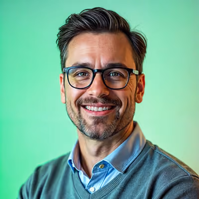 Smiling man with dark hair, beard, and glasses wearing a blue shirt under a gray sweater against a green gradient background.