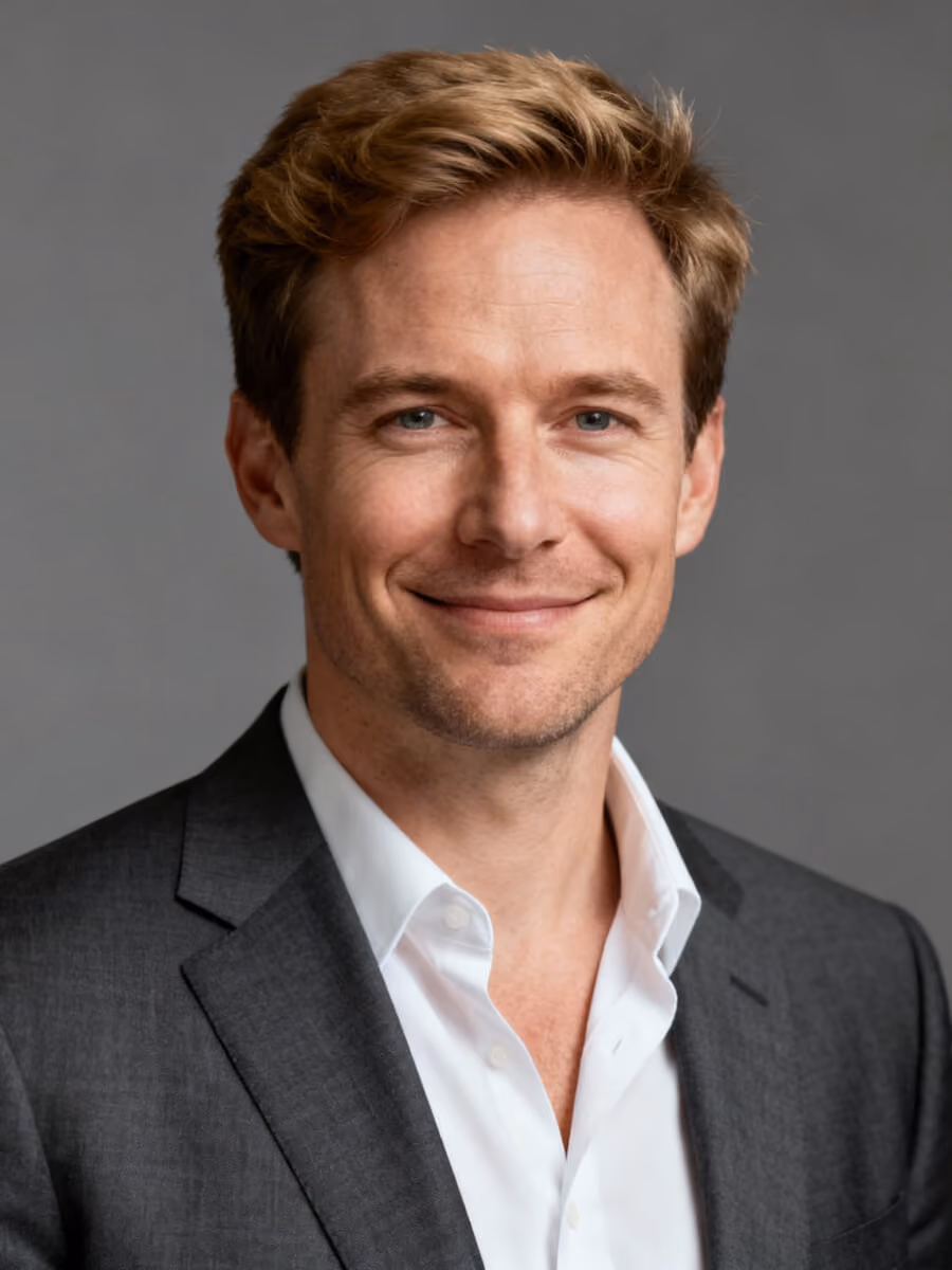 Portrait of a smiling man with short blonde hair wearing a dark gray suit jacket and white dress shirt against a gray background.