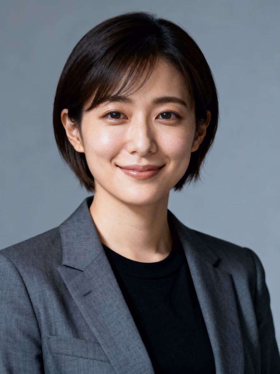 Smiling woman with short dark hair, wearing a gray blazer over a black top, against a plain gray background.