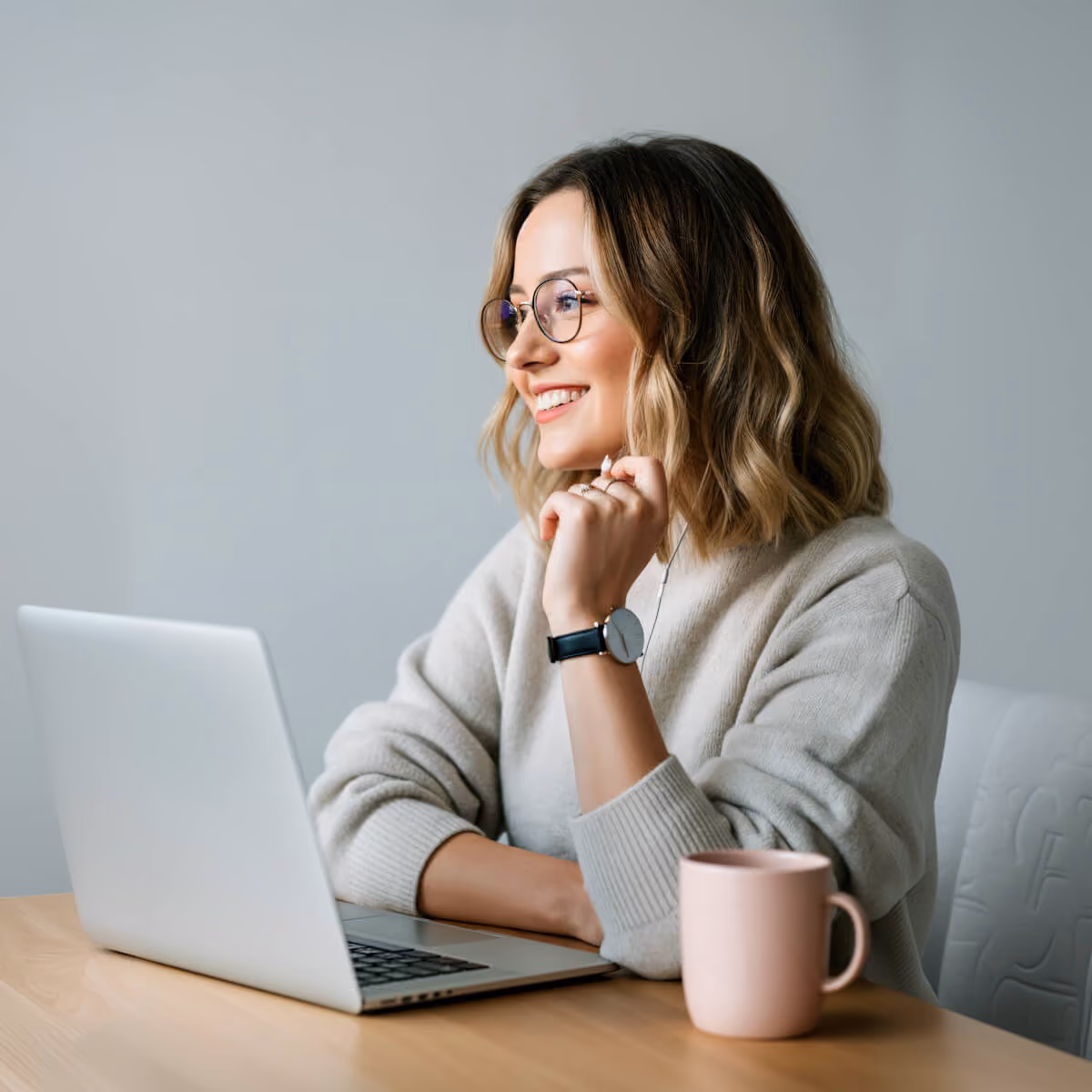 Smiling woman with glasses and wavy hair sitting at a wooden table using a laptop, with a pink mug nearby.