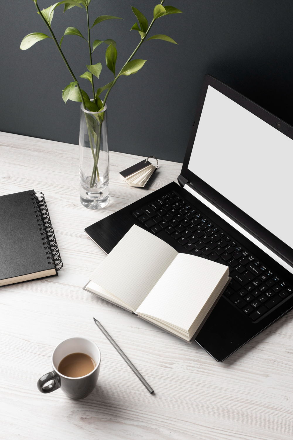 Minimal workspace with a black laptop, an open grid notebook on the keyboard, a closed spiral notebook, a vase with green leafy stems, a pencil, and a cup of coffee on a light wood desk against a dark background.