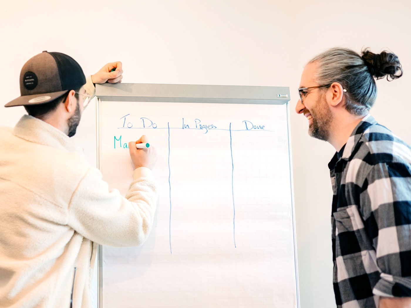 Dr. Nico Thümler schreibt an ein Flipchart mit den Spalten To Do, In Progress und Done, Moritz Keil schaut lachend zu