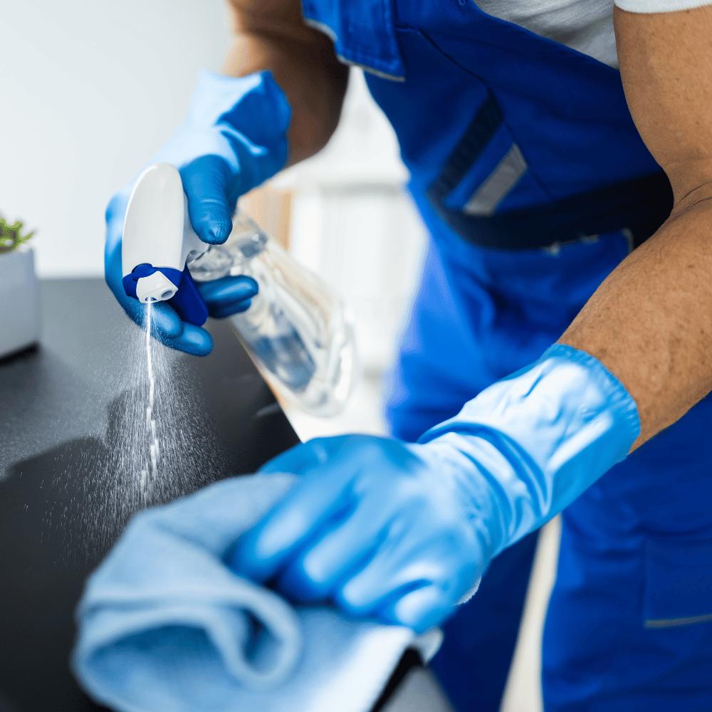 Reinigungskraft in blauer Arbeitskleidung sprüht Reiniger auf eine Arbeitsfläche