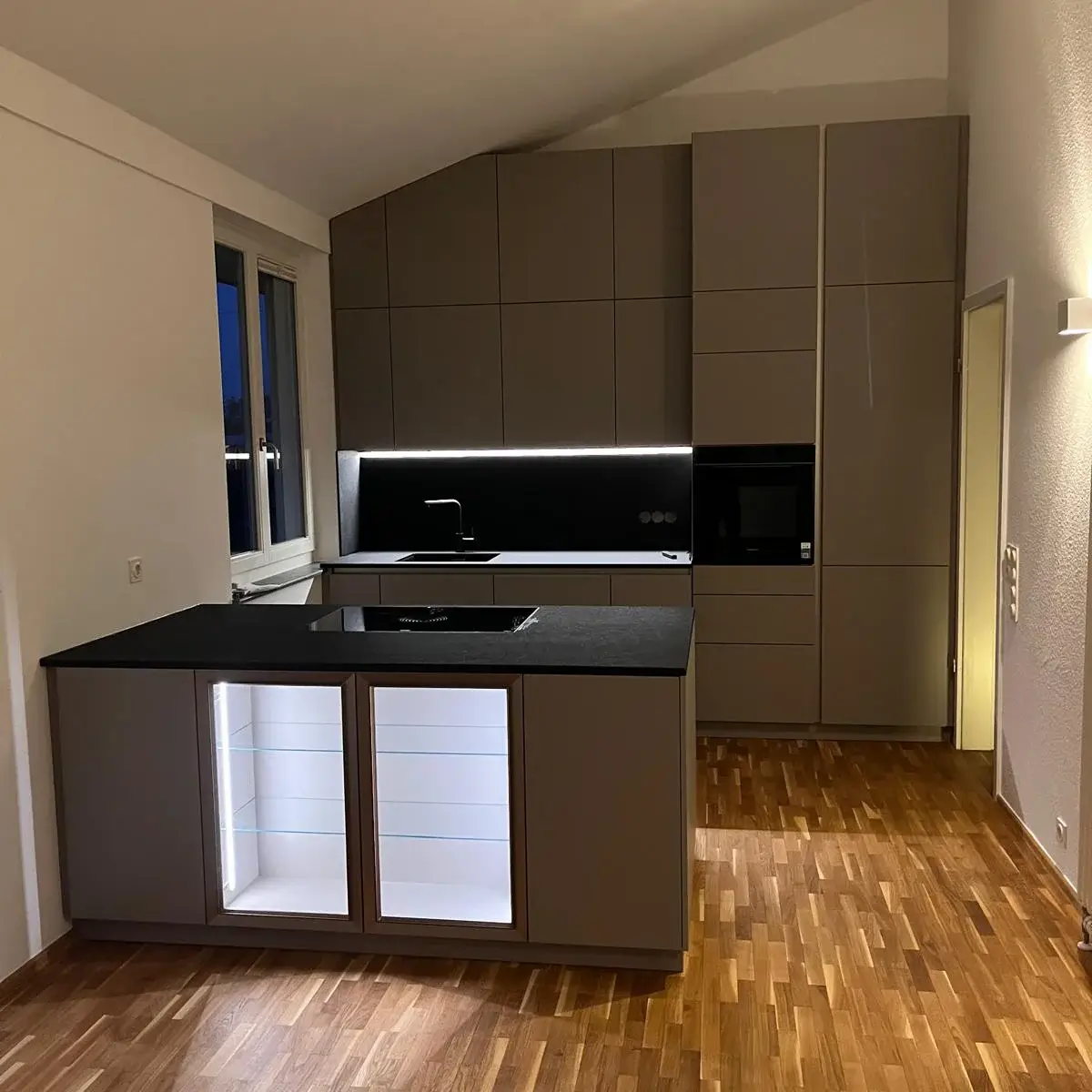 Moderne Küche mit grauen Schränken, schwarzer Arbeitsplatte und beleuchtetem Glasvitrinenschrank.