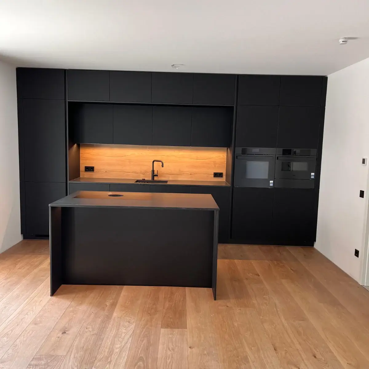 Moderne schwarze Küche mit Holzrückwand, Kücheninsel und hellbraunem Holzboden.