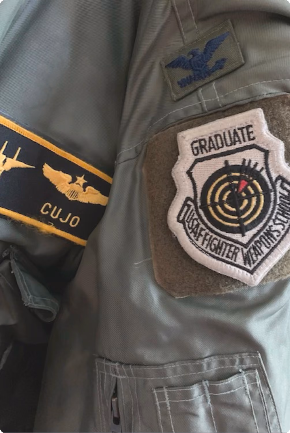 Close-up of Cujo's USAF flight suit with the Weapons School graduate patch, the fighter pilot background behind VMax Group's teamwork training