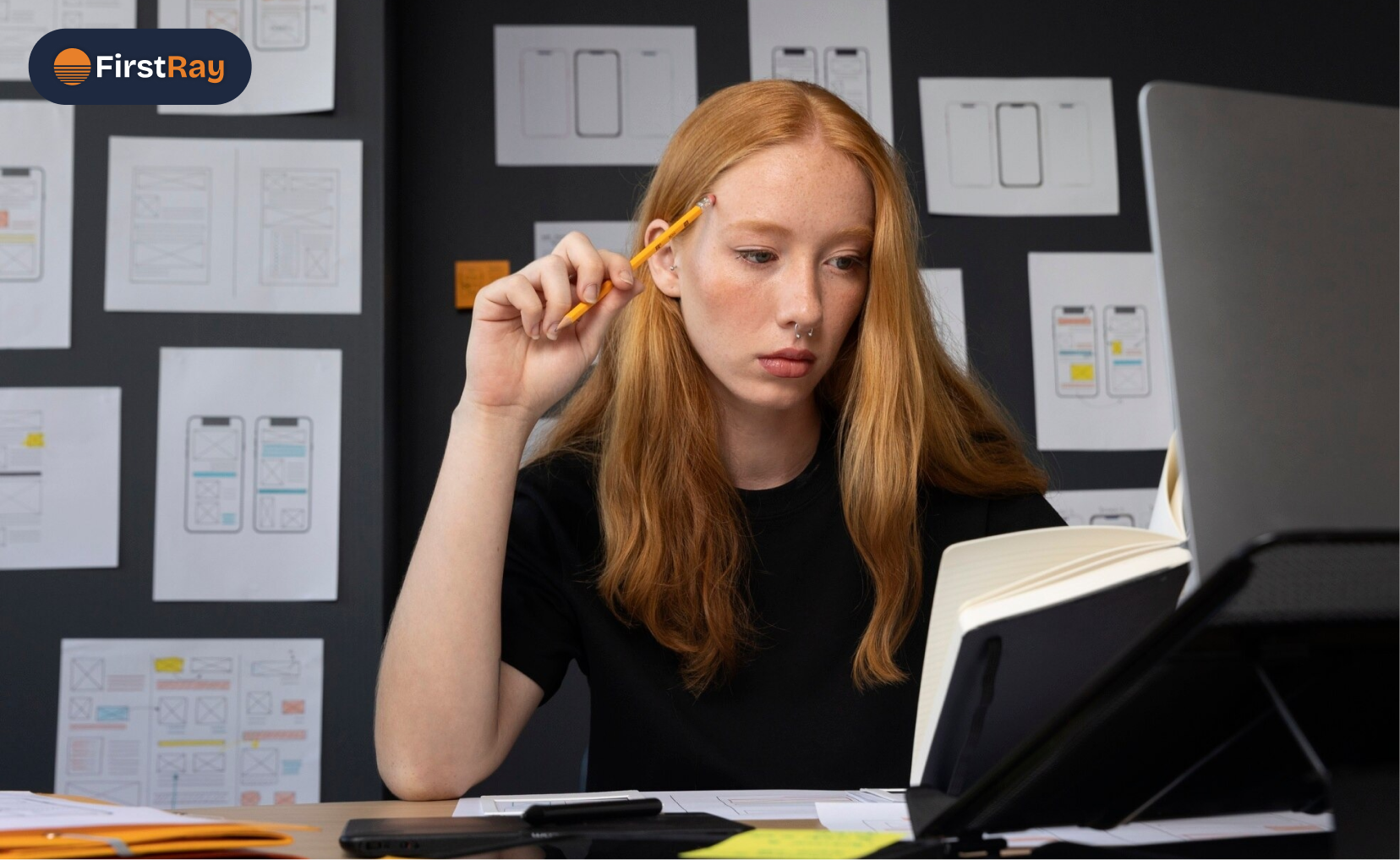 A focused young woman with long red hair and a black shirt holding a pencil thoughtfully to her temple while reading an open notebook, sitting at a desk with papers and a tablet, a wall behind her covered with design sketches and wireframes.
