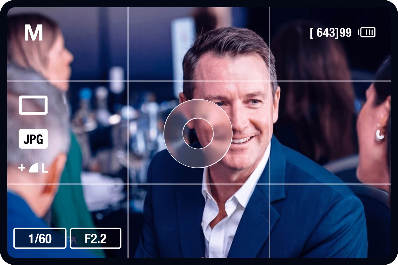 Viewfinder screen of a digital camera focused on a man in a blue suit smiling and talking to a woman in a social setting with blurred people and glasses in the background.