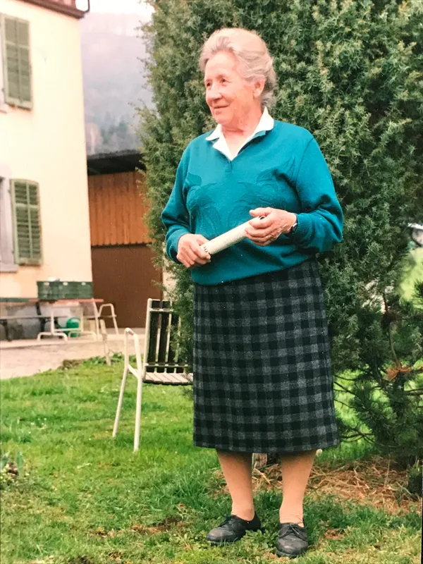 Lina Keller in türkisem Pullover und kariertem Rock vor dem Bauernhaus, einen Lauch in der Hand