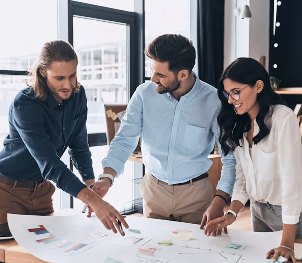 Trois collègues, deux hommes et une femme, collaborent autour d'une table de travail avec des graphiques et des notes adhésives, dans un bureau moderne avec de grandes fenêtres.