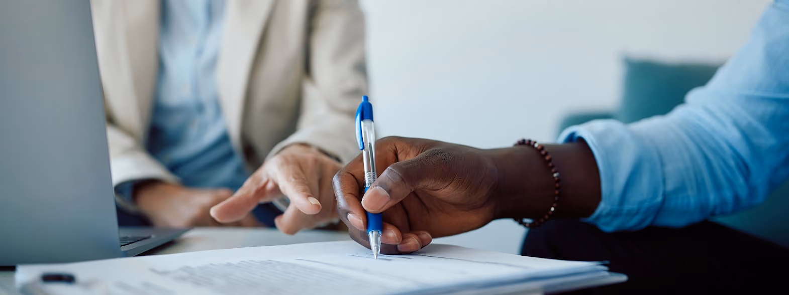 Une main tenant un stylo bleu écrivant sur des documents pendant qu'une autre main pointe vers ces documents, avec un ordinateur portable à côté.